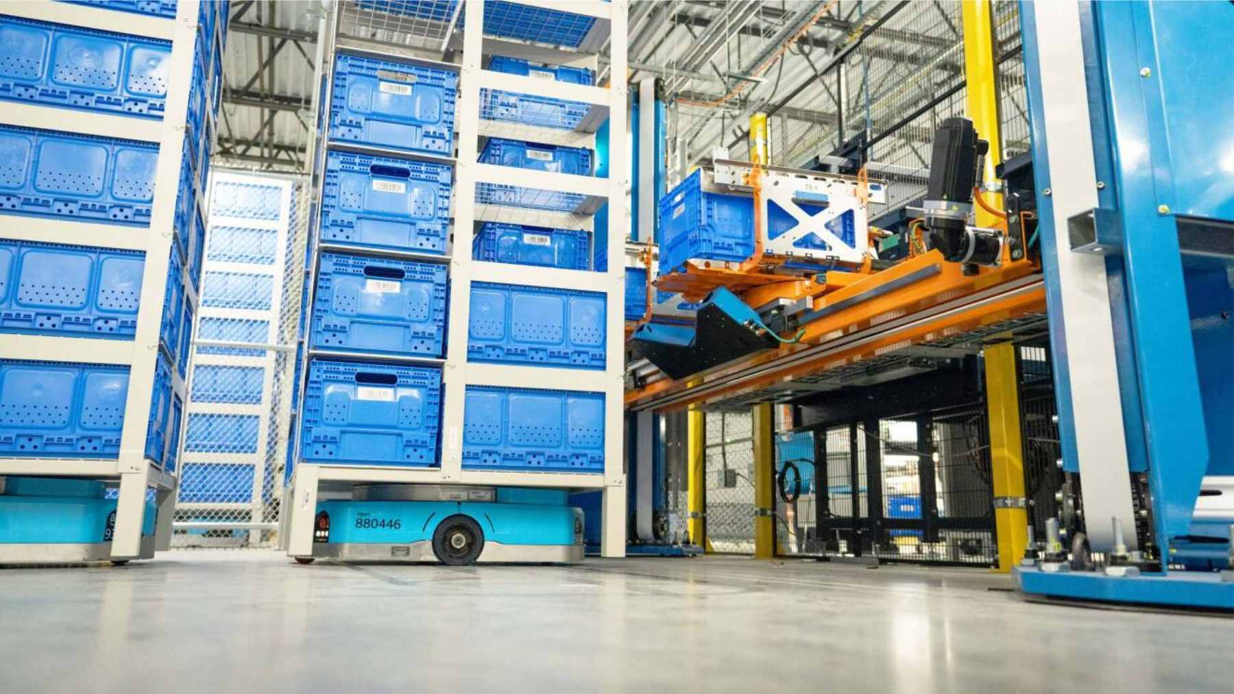 A light blue automated Amazon "Sequoia" mobile robot lifting and moving a tall metal shelving rack filled with blue storage totes in a fulfillment center.