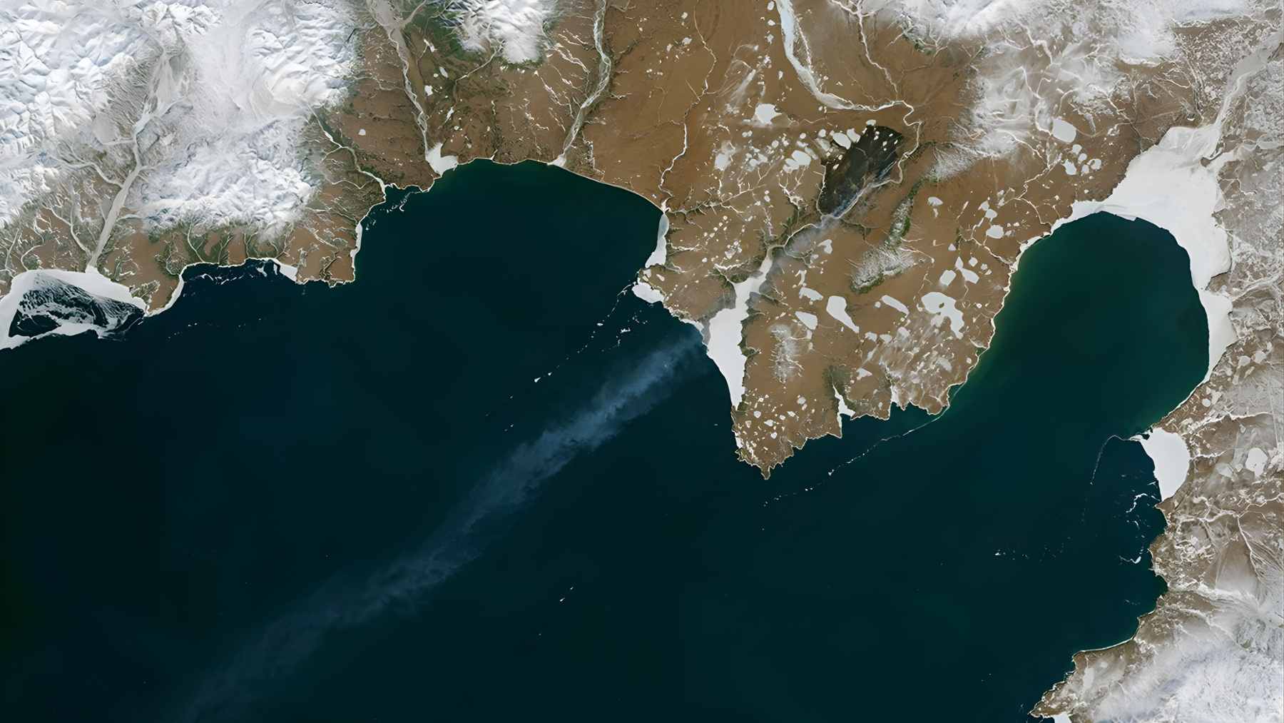 Satellite view of dark wildfire smoke plumes drifting over white snow-covered tundra and frozen lakes in the Arctic.