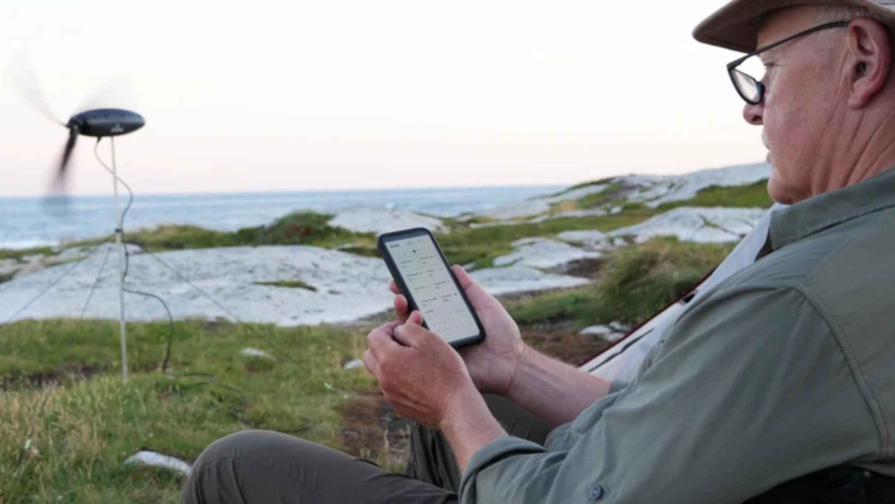 Person monitoring the Shine 2 portable wind turbine on a smartphone while using the device for off-grid power near the coast.