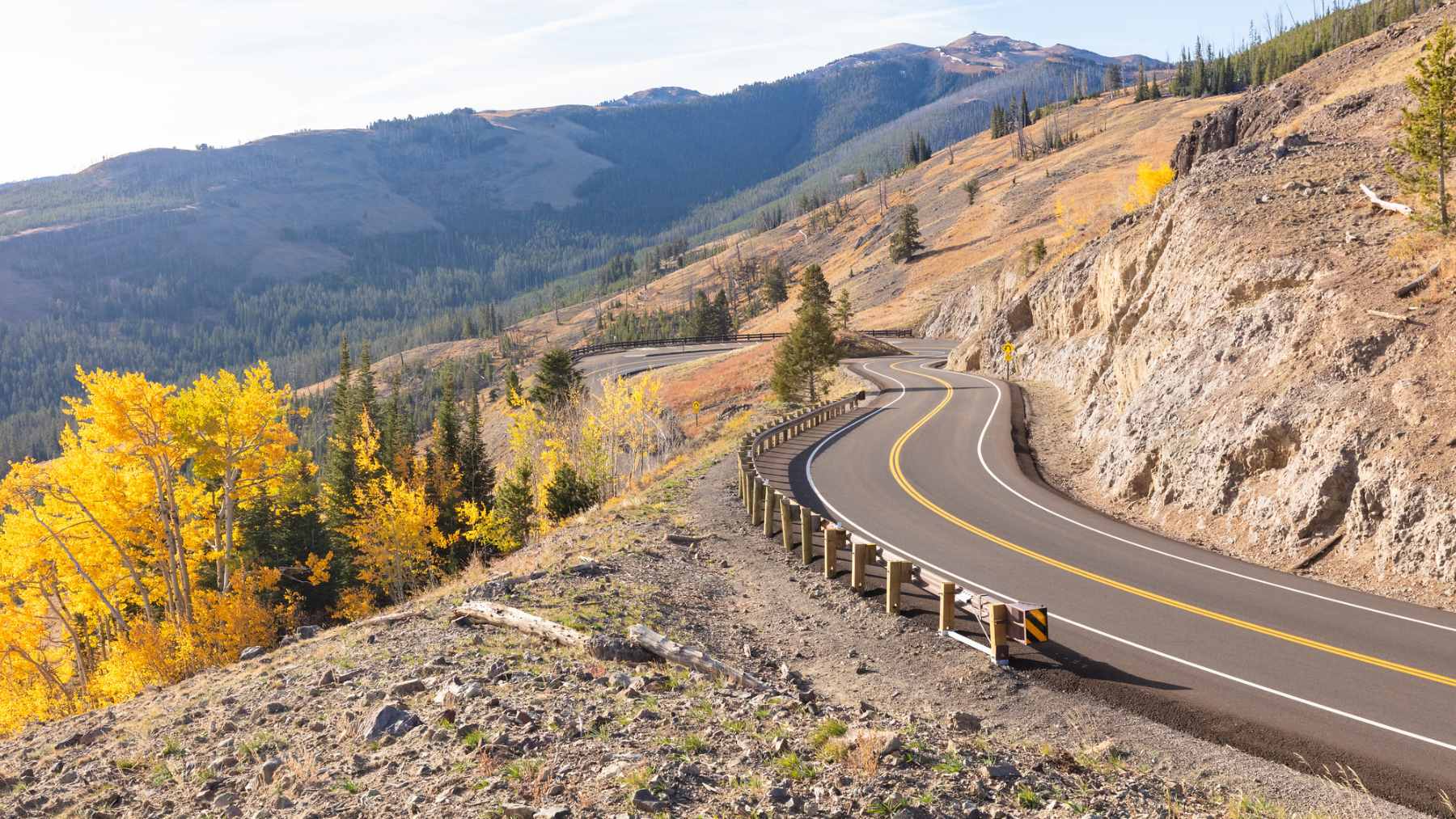 A winding, high-altitude paved road along the Beartooth Highway, featuring sweeping views of alpine tundra and distant snow-capped mountains.
