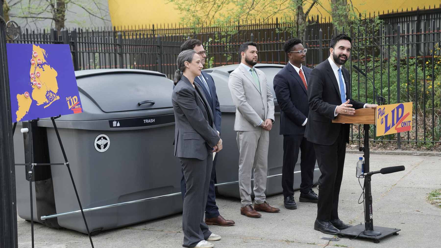 A line of locked, grey Empire Bins taking up a parking spot on a busy New York City street.