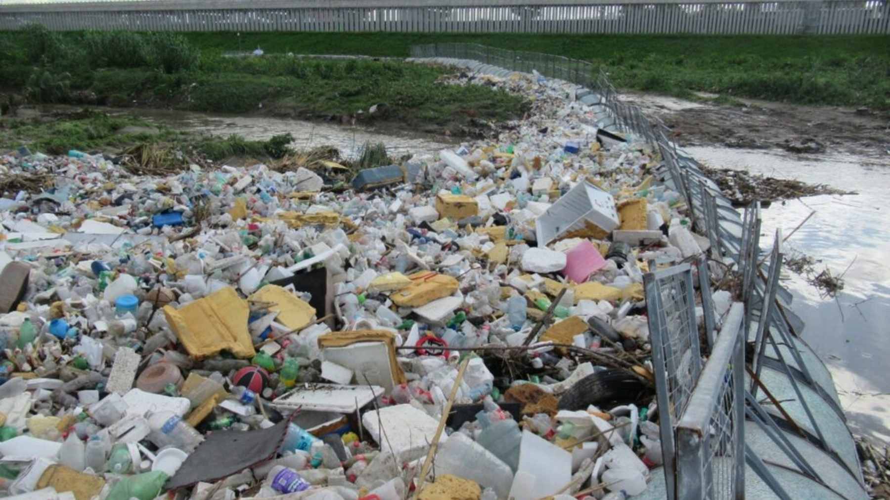 A heavy-duty floating trash boom spanning the concrete Tijuana River Flood Control Channel, trapping a large accumulation of plastic waste and debris.
