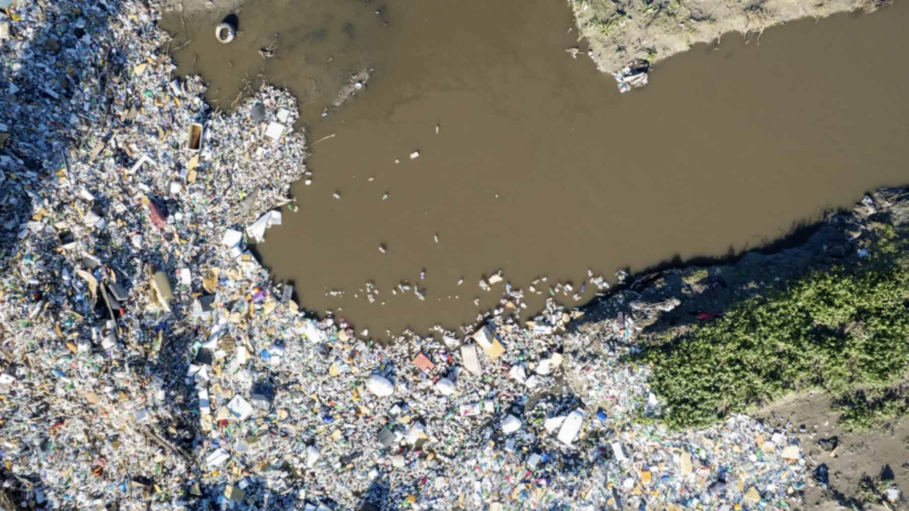 A heavy-duty floating trash boom spanning the concrete Tijuana River Flood Control Channel, trapping a large accumulation of plastic waste and debris.